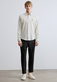 Dryžuoti marškiniai su sagutėmis šviesiai kreminės spalvos, su mažyčiu išsiuvinėtu logotipu; derinami su juodomis kelnėmis ir baltais sportbačiais.