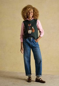 Kootud roheline vest, millel on rebase motiiv, seljas heledat roosat pikkade varrukatega särki, koos siniste teksapükste ja pruunide kingadega, seistes beežil pinnal.