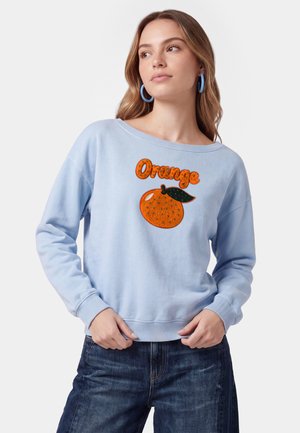 Ung kvinde iført lyseblå sweatshirt med teksten "Orange" og orange frugtmotiv, kombineret med mørkeblå jeans og blå øreringe.