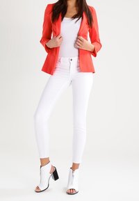 Blazer rosso aderente, canotta bianca, jeans skinny bianchi e stivaletti alla caviglia aperti bianchi con cerniera. Tessuto liscio, design sartoriale.