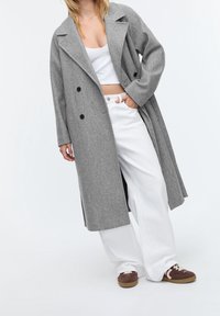 Femme portant un long manteau gris croisé, un débardeur blanc court, un pantalon large blanc et des baskets marron, debout avec une main dans la poche.