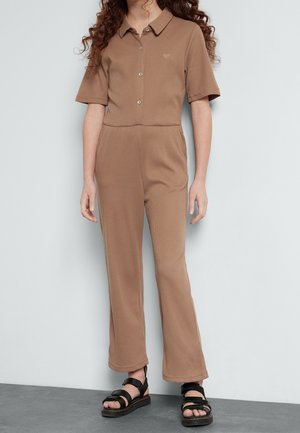 Personne portant une combinaison marron à manches courtes avec boutons, avec un petit logo en forme de cœur sur la poitrine, associée à des sandales noires.