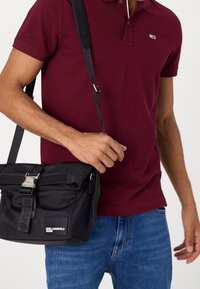 Black nylon shoulder bag with a flap closure and metal buckle. Features logo patch, adjustable strap, and structured shape.
