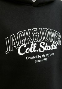 Melns kapuce ar baltu tekstu "JACK & JONES Coll. Studio Created by the J&J crew Since 1990" uz krūšu daļas.