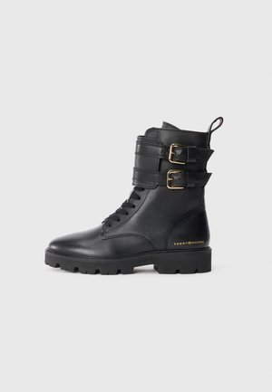 Black leather ankle boots featuring two buckle accents, a lace-up front, and textured rubber sole for grip. Gold logo on the side.