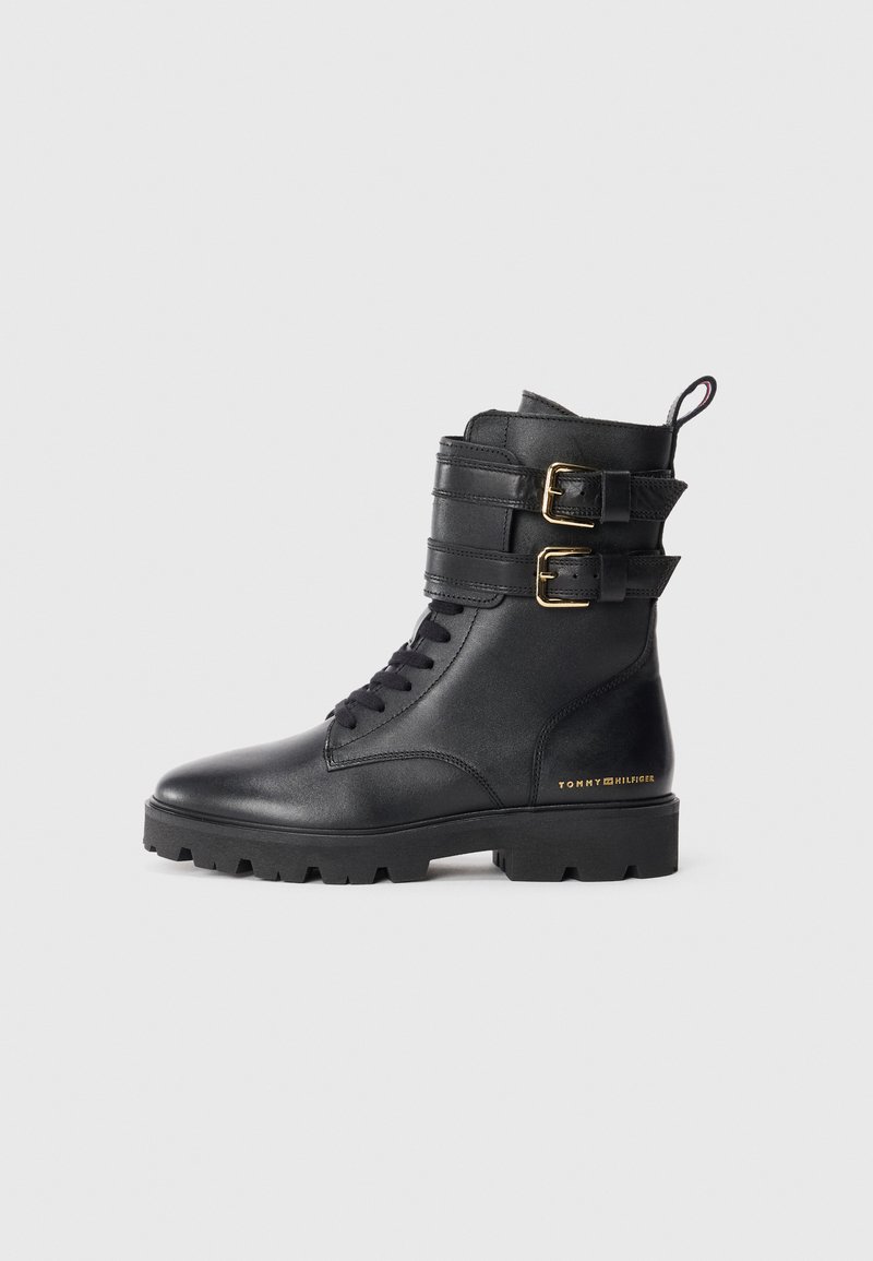 Botas de tornozelo em couro preto com dois detalhes de fivela, atacadores à frente e sola de borracha texturizada para aderência. Logótipo dourado na lateral.