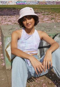 Top crop em tie-dye roxo claro com detalhe em nó, combinado com jeans de perna larga azul claro, e um chapéu de balde pálido com um logotipo.