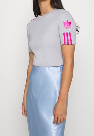 Femme portant une chemise à manches courtes gris clair avec des rayures roses et un logo sur la manche, associée à une jupe en satin bleu clair.