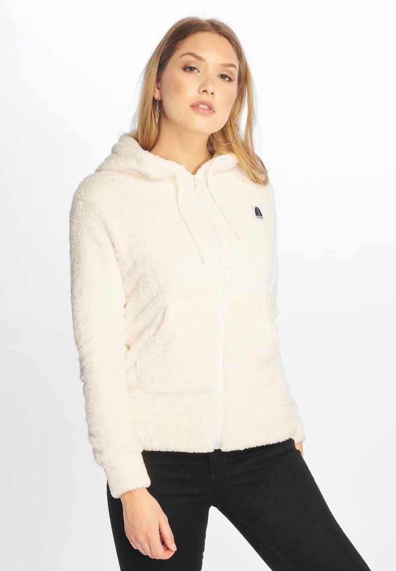 Sudadera blanca de forro polar con cremallera, capucha y bolsillos frontales, que presenta una textura suave y un pequeño logo en el lado izquierdo del pecho. Se llevan pantalones negros.