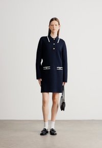 Tengerészkék kötött ruha galléros nyakkivágással, fehér szegéllyel és két csíkos zsebbel. A modell fekete cipőt visel és egy fekete kézitáskát tart.