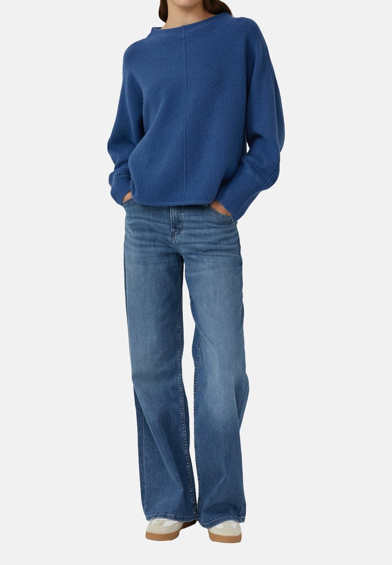 Femme portant un pull bleu à manches longues, un jean bleu à jambes larges et des chaussures blanches, debout les mains dans les poches.