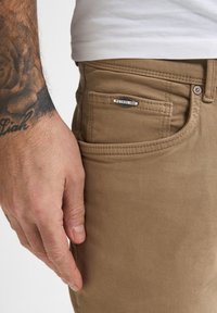 Pantalones de algodón en color marrón claro que presentan un bolsillo frontal con una etiqueta metálica "PETROLIND", exhibidos junto a una mano con tatuajes visibles.
