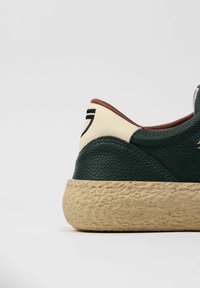 Sneaker verde in pelle con superficie texturizzata, tallone color crema e intersuola in juta intrecciata. Presenta un marchio nero sul tallone.