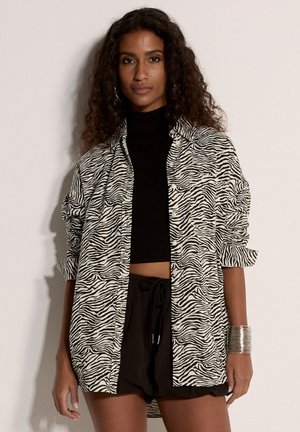 Mujer con cabello rizado que lleva un crop top negro, una camisa oversized con estampado de cebra, pantalones cortos negros y una pulsera ancha plateada, posando frente a un fondo liso.