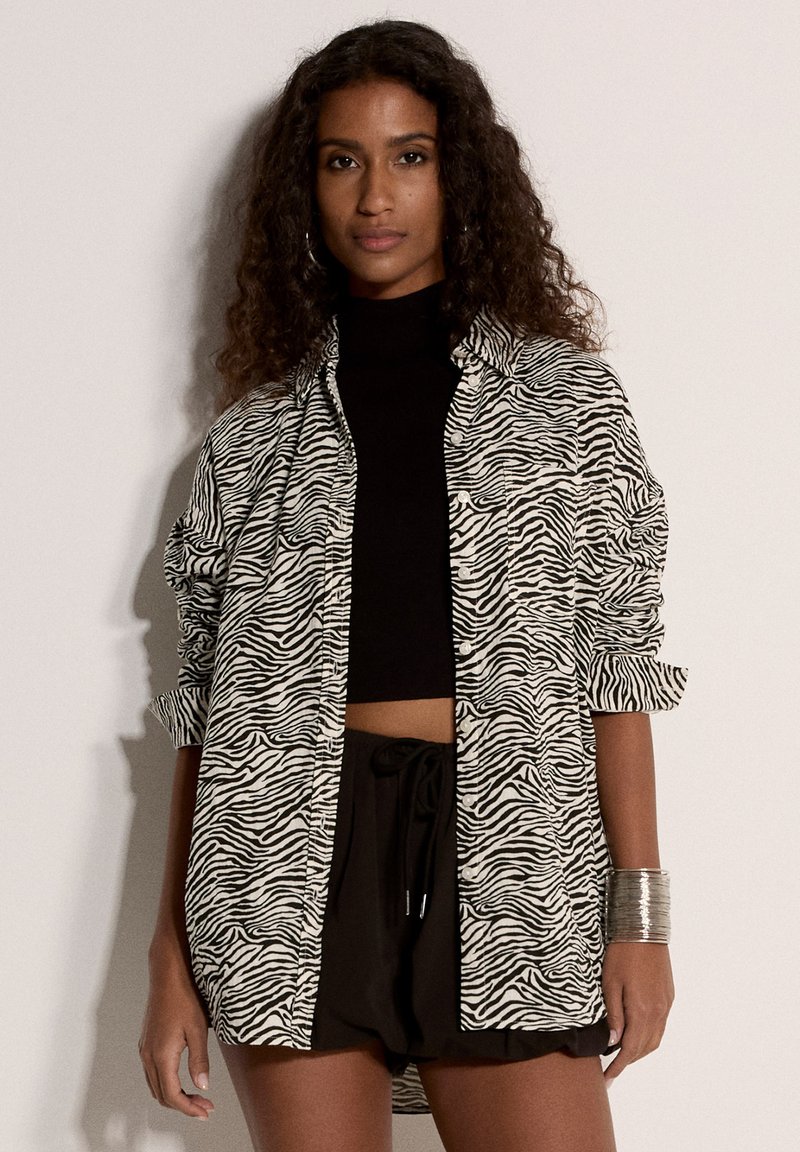Femme aux cheveux bouclés portant un crop top noir, une chemise oversize à motif zèbre, un short noir et un bracelet manchette argenté, debout devant un fond uni.
