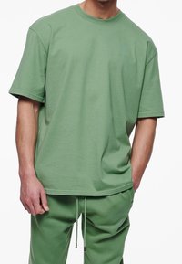 Zelené bavlněné tričko s volným střihem, kulatým výstřihem, krátkými rukávy a jemným logem na hrudi. Hladká, jednobarevná textura.