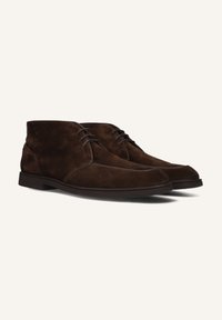 Bottines en daim marron avec un bout arrondi et des coutures lisses, présentant un design à lacets et une semelle plate en caoutchouc.