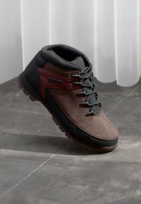 Brown suede hiking shoe with black rubber sole, grey laces, and maroon accents. Features a padded collar and a textured exterior.