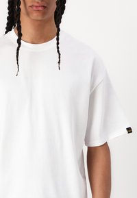 T-shirt oversize blanc en coton à manches courtes, avec une étiquette noire sur la manche gauche et une coupe décontractée. Texture lisse.