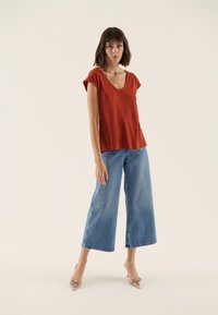 Rostkleurig kort mouwtopje met een diepe V-hals; gecombineerd met wijde blauwe spijkerbroeken en doorzichtige schoenen met een puntige neus.