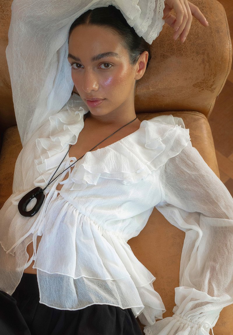 Blusa blanca con volantes y tejido transparente y de mangas largas. Collar con colgante circular negro, llevado sobre una prenda negra en capas. Fondo de cuero marrón.