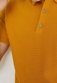 Short-sleeved polo shirt in mustard color, features a textured striped pattern, three-button placket with gray buttons, and ribbed collar.