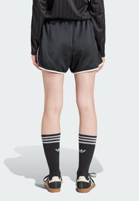 Černé saténové sportovní šortky s bílým lemem, nošené s černými vysokými ponožkami do kolen, které mají tři pruhy a logo Adidas, a černými teniskami.