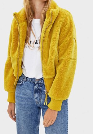 Person wearing a bright yellow fleece zip-up jacket, white graphic t-shirt, and blue high-waisted jeans against a plain background.