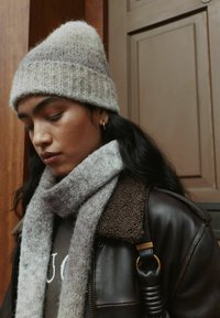 Light gray knitted beanie with ribbed texture, paired with a soft gray scarf. Worn with a brown leather jacket featuring a fur collar.
