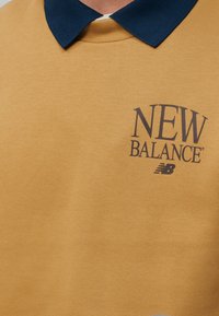 Φούτερ μουσταρδί με σκούρο μπλε γιακά, με μαύρο λογότυπο «New Balance» στο στήθος.