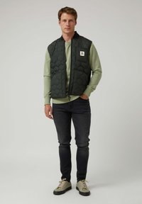 Chaleco acolchado de color verde oscuro con patrón acolchado, cierre de cremallera y bolsillos laterales, llevado sobre una sudadera verde claro y combinado con vaqueros oscuros.