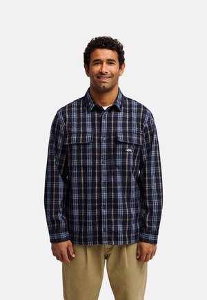Homme souriant portant une chemise à carreaux bleu marine et marron avec des poches avant et un pantalon kaki sur un fond uni.
