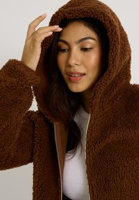Veste à capuche marron et duveteuse avec un zip à l'avant, caractérisée par une texture douce et une capuche doublée. Modèle touchant ses cheveux, portant un haut blanc.