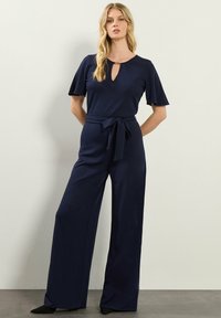 Tuta navy con maniche corte a volant, scollatura a chiave, vita con cintura e pantaloni a gamba larga, realizzata in tessuto morbido.