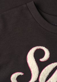 Gros plan sur un tissu foncé avec un col côtelé et une lettre S blanche texturée bordée d'une couture rose sur le vêtement.