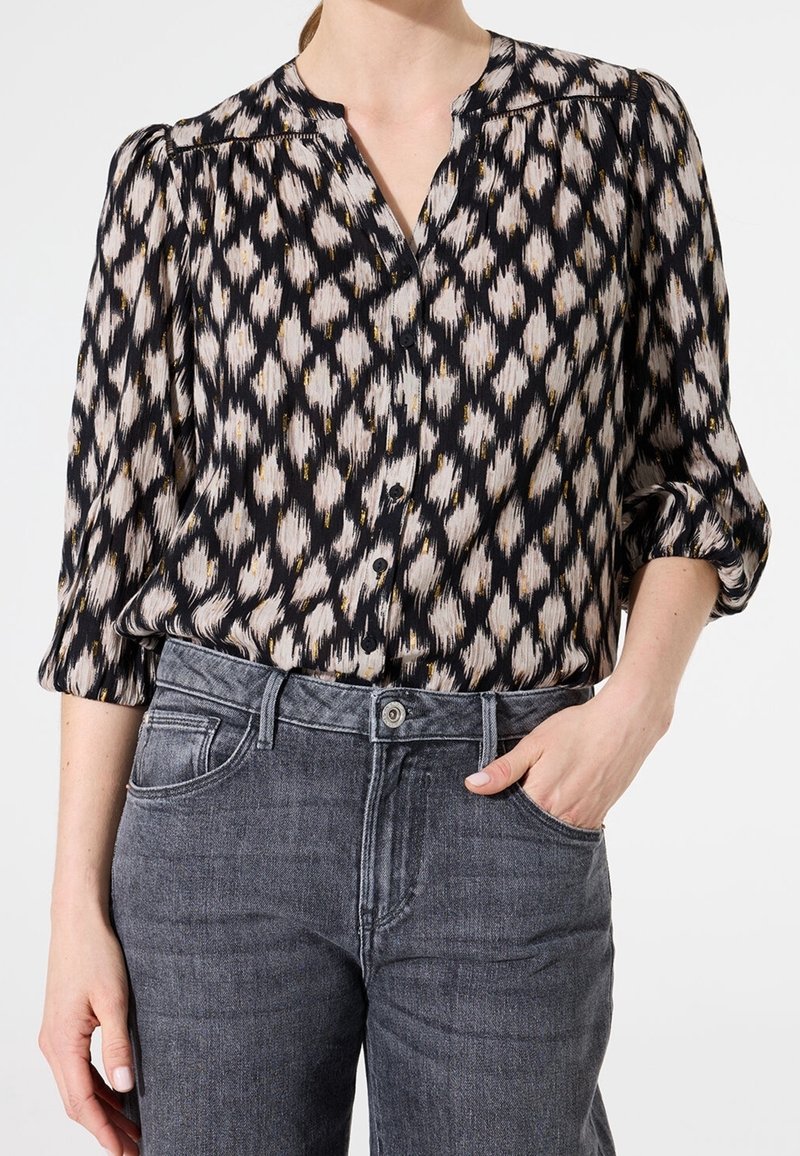 Femme portant une blouse boutonnée à motifs noirs et beiges avec des manches trois-quarts et un jean gris taille haute, main dans la poche.