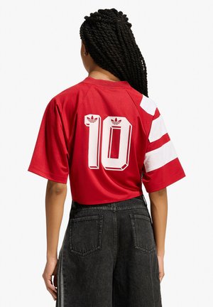 Persona con lunghi capelli intrecciati che indossa una maglia sportiva rossa con il numero 10 e jeans neri a vita alta, in piedi di spalle.