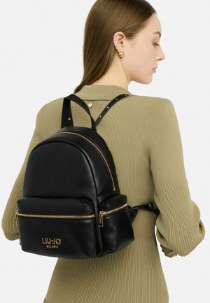 Zaino in pelle nera con silhouette arrotondata, dettagli in metallo dorato e chiusura superiore a zip. Presenta una tasca anteriore con logo. Spallacci regolabili.