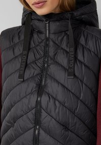 Frau in einer schwarzen gesteppte, zip-up Kapuzenweste mit schwarzen Kordeln, grauem Text auf den Kordeln und einem burgunderfarbenen Langarmshirt darunter.