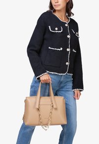 Beige håndtaske med to håndtag og kædeaccent, lavet af glat materiale. Har et struktureret design og minimalistisk branding.