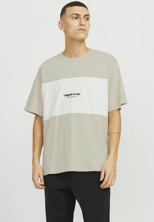Camiseta de manga corta de color beige claro con una franja horizontal blanca y el logo en texto negro "Originals Studio" en el pecho. Tejido de algodón suave.