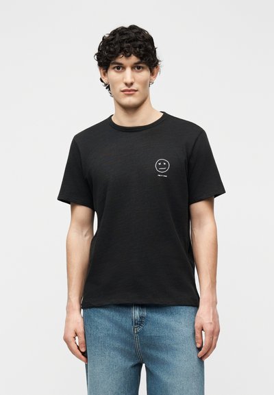Man with curly hair wearing black t-shirt with small white crossed-eyes smiley face and text on chest, paired with blue jeans.