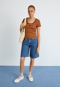 Haut ajusté rayé marron et orange avec un col rond, porté avec un short en denim bleu et des chaussures blanches. Le mannequin tient un sac en tricot.