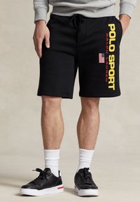 Schwarze Shorts mit einer weichen Textur, auf denen in auffälligem Gelb der Schriftzug "POLO SPORT" und eine amerikanische Flagge auf dem linken Oberschenkel gedruckt sind. Kombiniert mit schwarzen Sneakers.