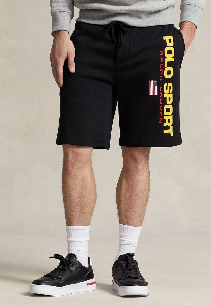 Schwarze Shorts mit einer weichen Textur, auf denen in auffälligem Gelb der Schriftzug "POLO SPORT" und eine amerikanische Flagge auf dem linken Oberschenkel gedruckt sind. Kombiniert mit schwarzen Sneakers.