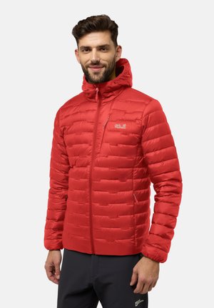 Mann mit kurzen dunklen Haaren, der eine rote gesteppte Jacke mit Reißverschluss und Kapuze sowie schwarze Hose vor schlichtem Hintergrund trägt.
