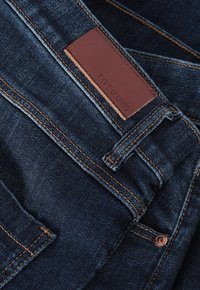 Dark blue denim jeans with copper stitching, featuring a brown leather patch embossed with "TNTUUM" and standard button closure.
