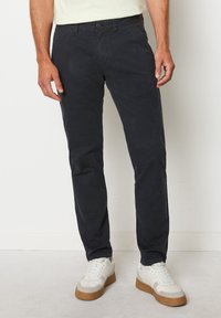 Chino navy in misto cotone, design a gamba dritta, con tasche frontali e posteriori, abbinati a sneaker bianche e sfondo chiaro.