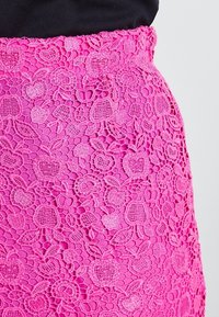 Světle růžová krajková sukně s květinovými a ovocnými vzory nošená s černým topem, ukazující podrobné vyšívání a texturu.