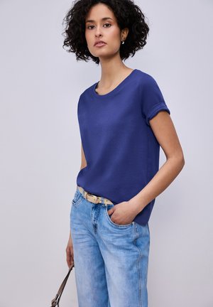 Mujer con cabello rizado que lleva una camiseta azul con las mangas remangadas y jeans azul claro, sosteniendo el asa de una bolsa con una mano y con la otra en el bolsillo.